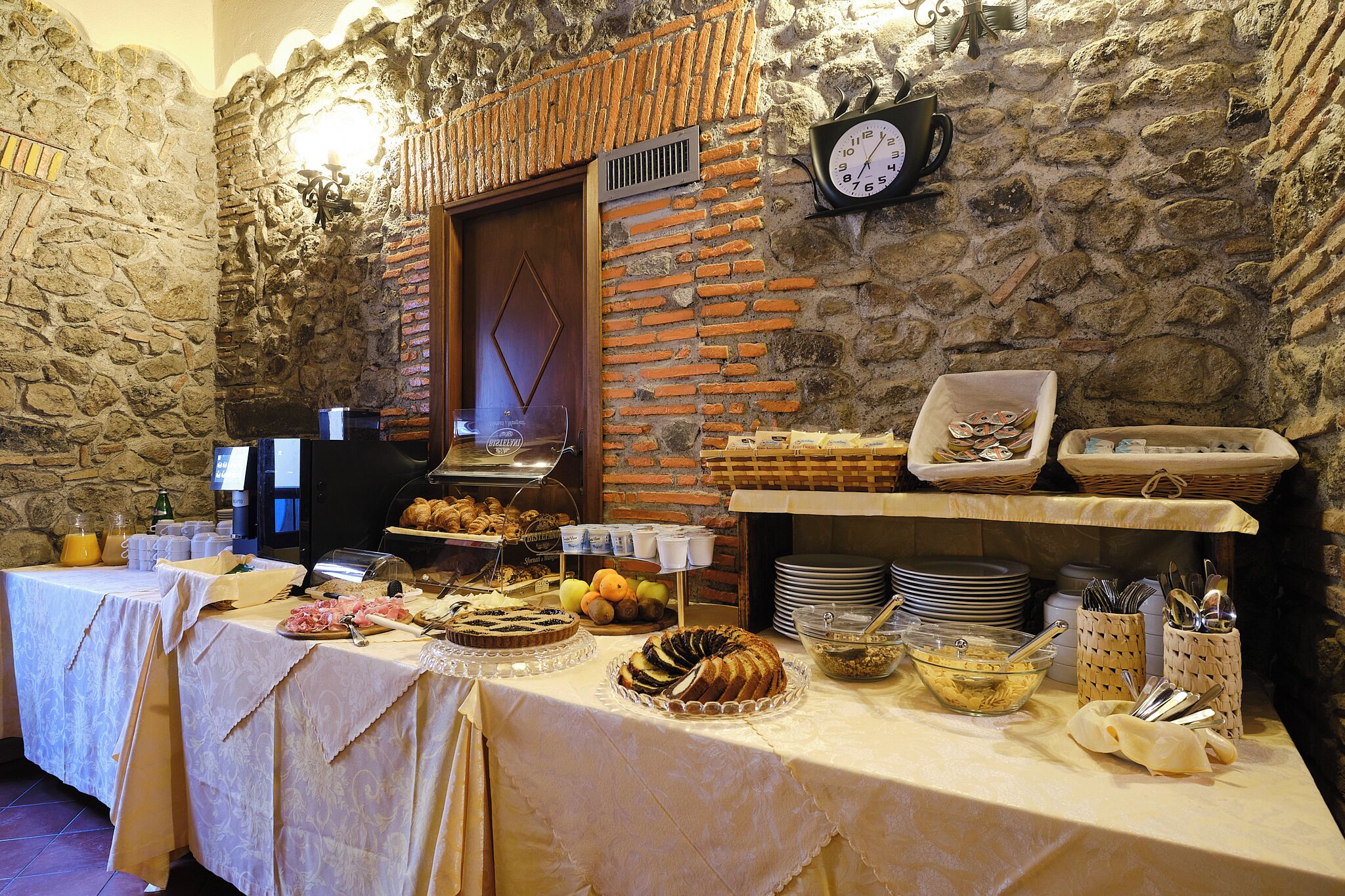 Una mesa de buffet con diferentes alimentos y utensilios, frente a una pared de piedra con un reloj.
