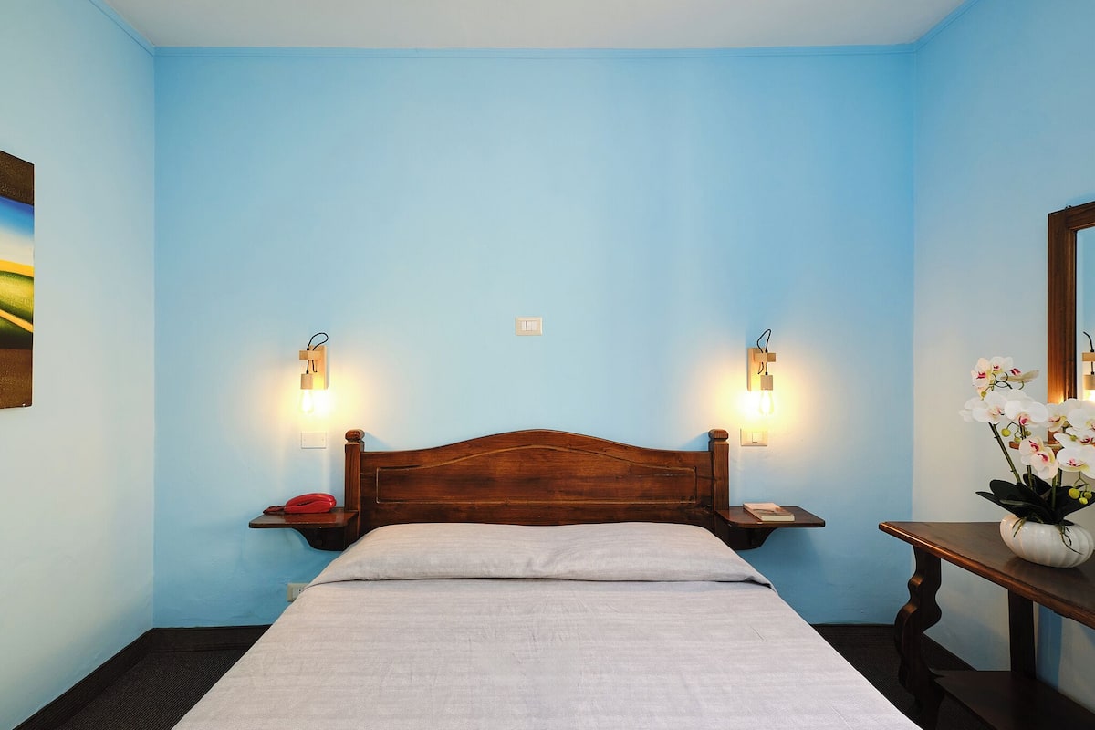 A hotel room with a wooden headboard, two bedside tables with lamps, a bed with a white sheet, and a small table with a vase of flowers.
