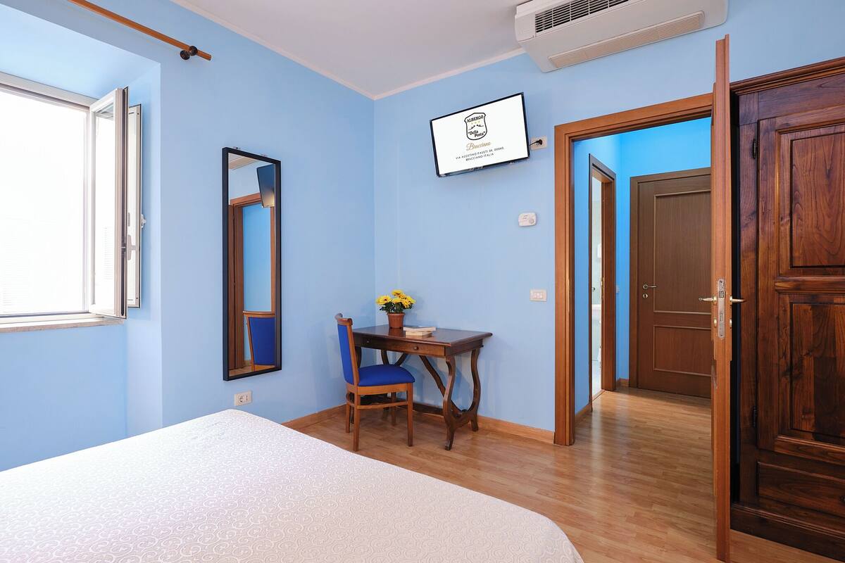 A bedroom with a bed, a desk with a chair, a mirror, and a door leading to another room.