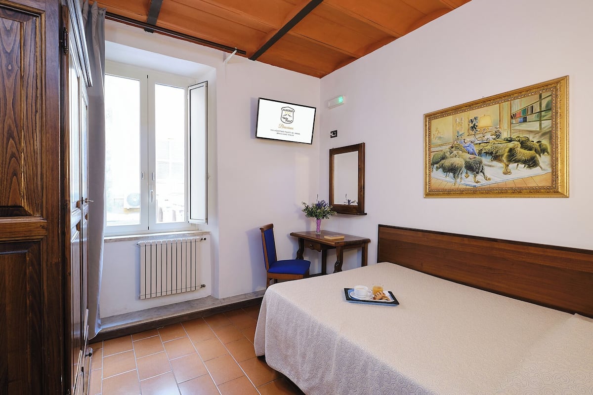 A hotel room with a bed, a desk with a chair, a window, a radiator, and a painting on the wall.