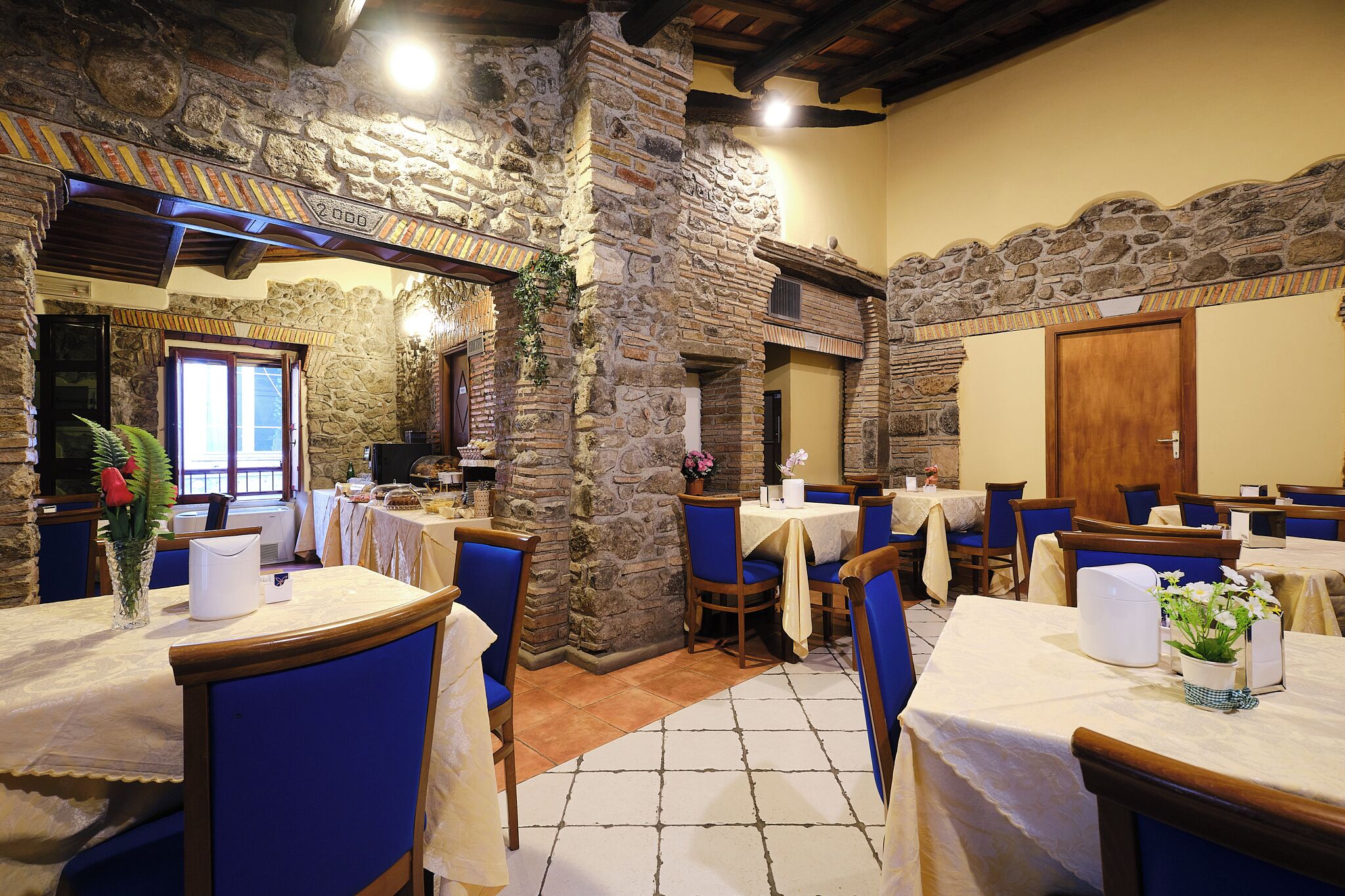 Un restaurant aux murs de pierre, aux poutres apparentes et aux tables recouvertes de nappes blanches, agrémentées de chaises bleues.