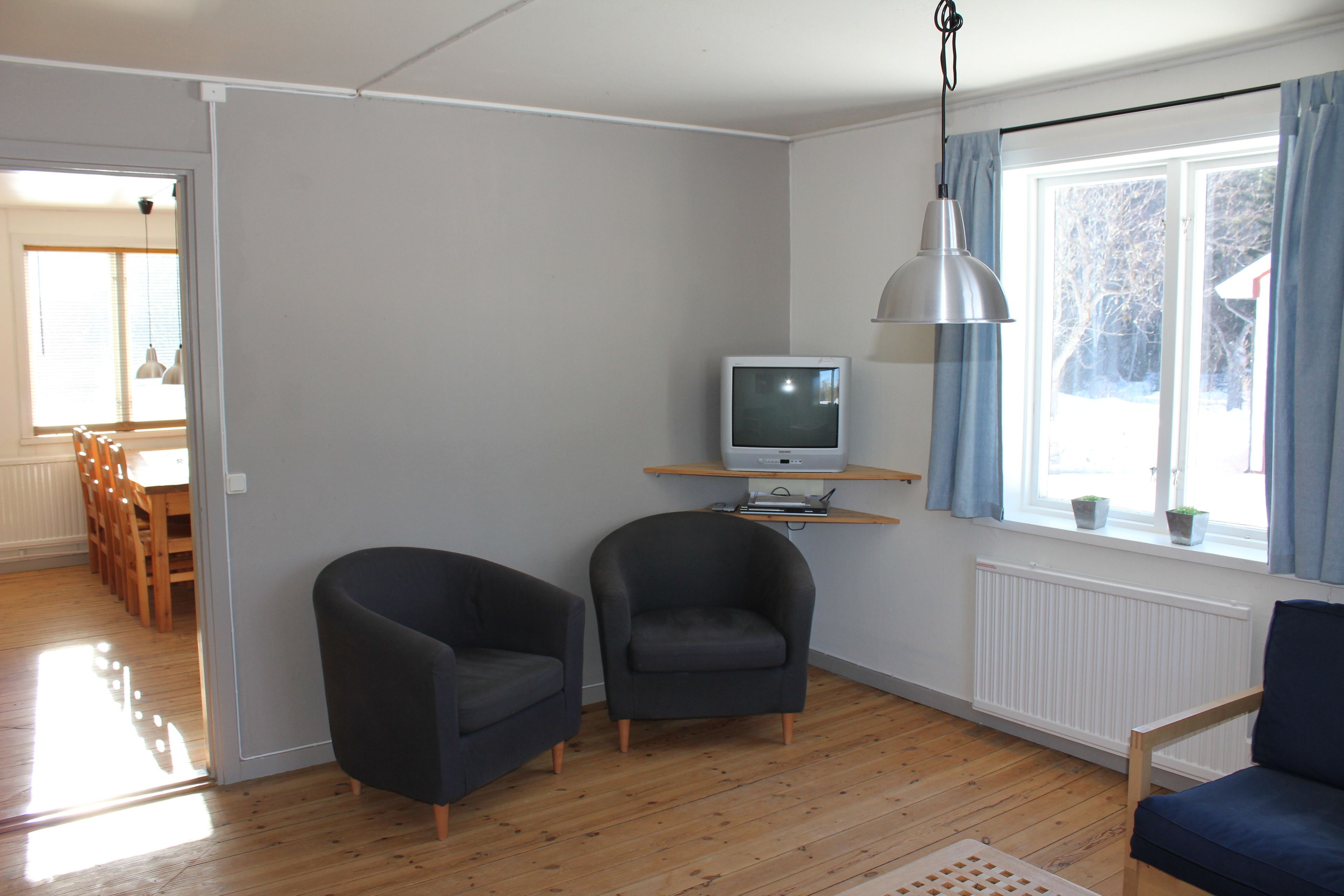 Una sala de estar con dos sillones, un televisor sobre una estantería, un ventanal con cortinas y piso de madera.