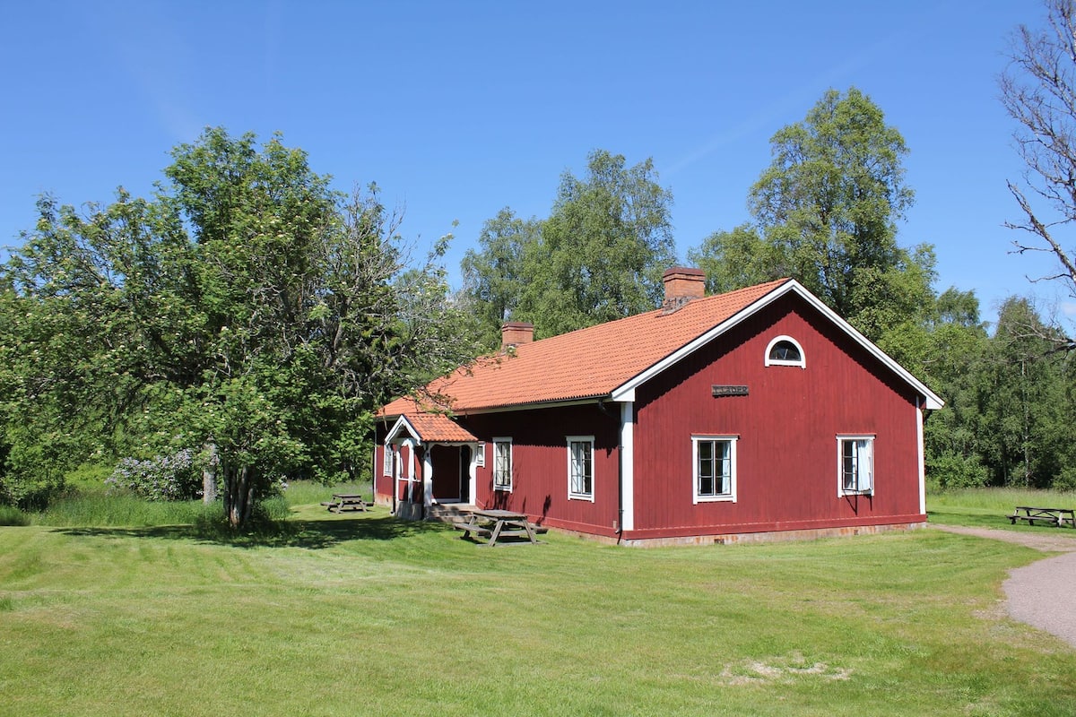 A red wooden building with a sloped roof, surrounded by green grass and trees.