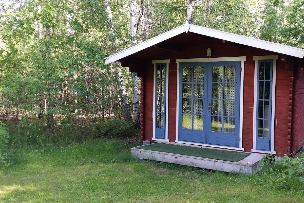 A small wooden cabin with a red exterior and blue doors, surrounded by greenery.