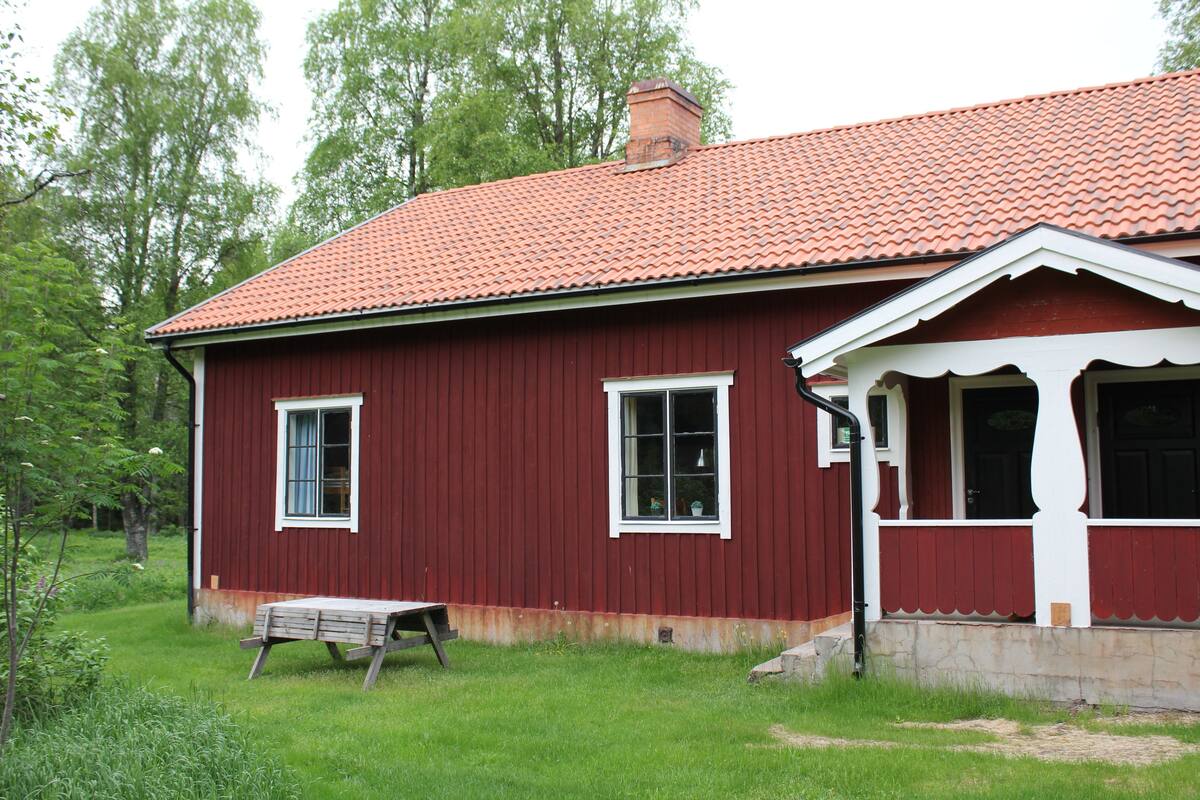 A red wooden house with a tiled roof, a porch, and a bench in the yard.