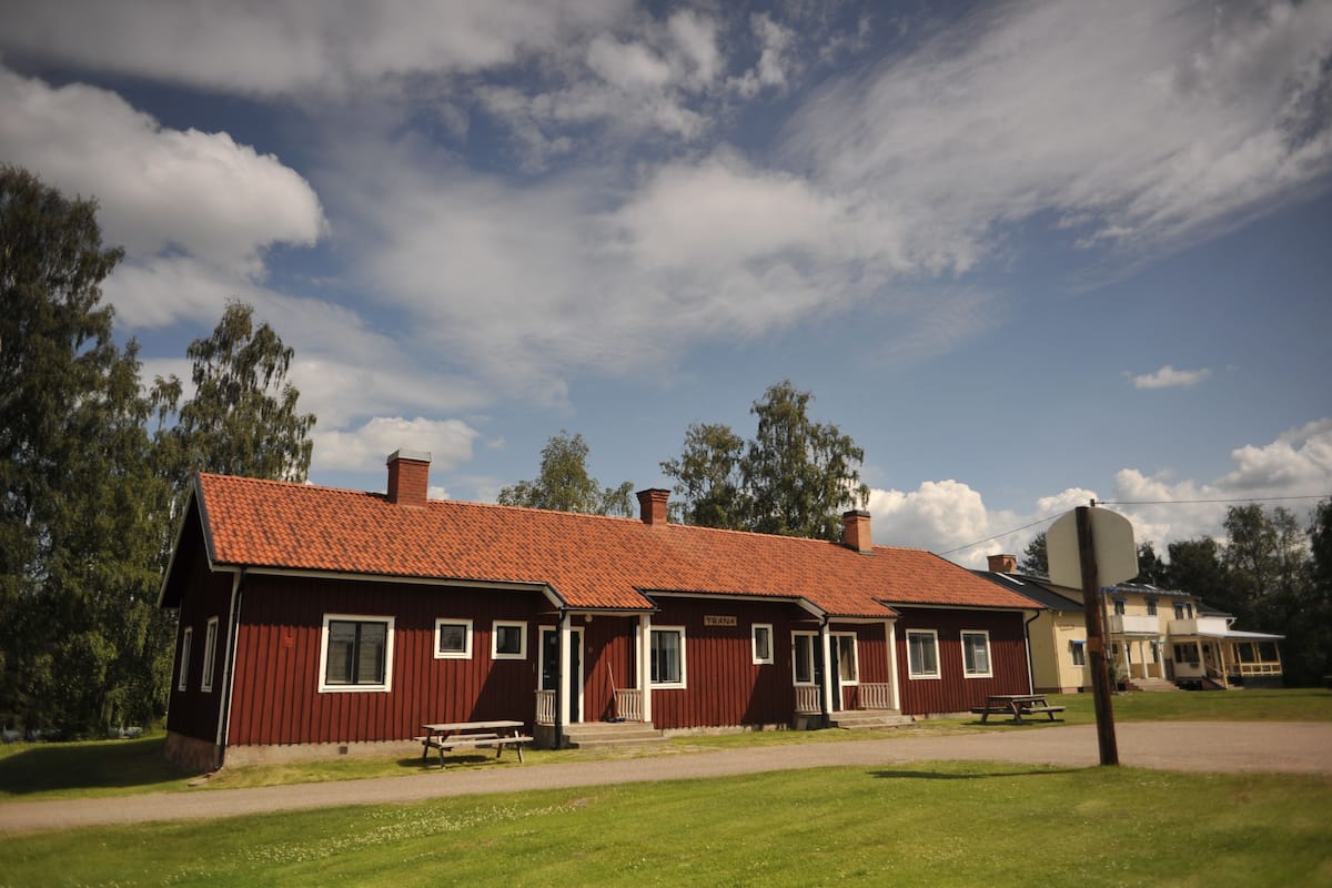 A red-roofed building with a bench and picnic table in front, surrounded by green grass and trees.