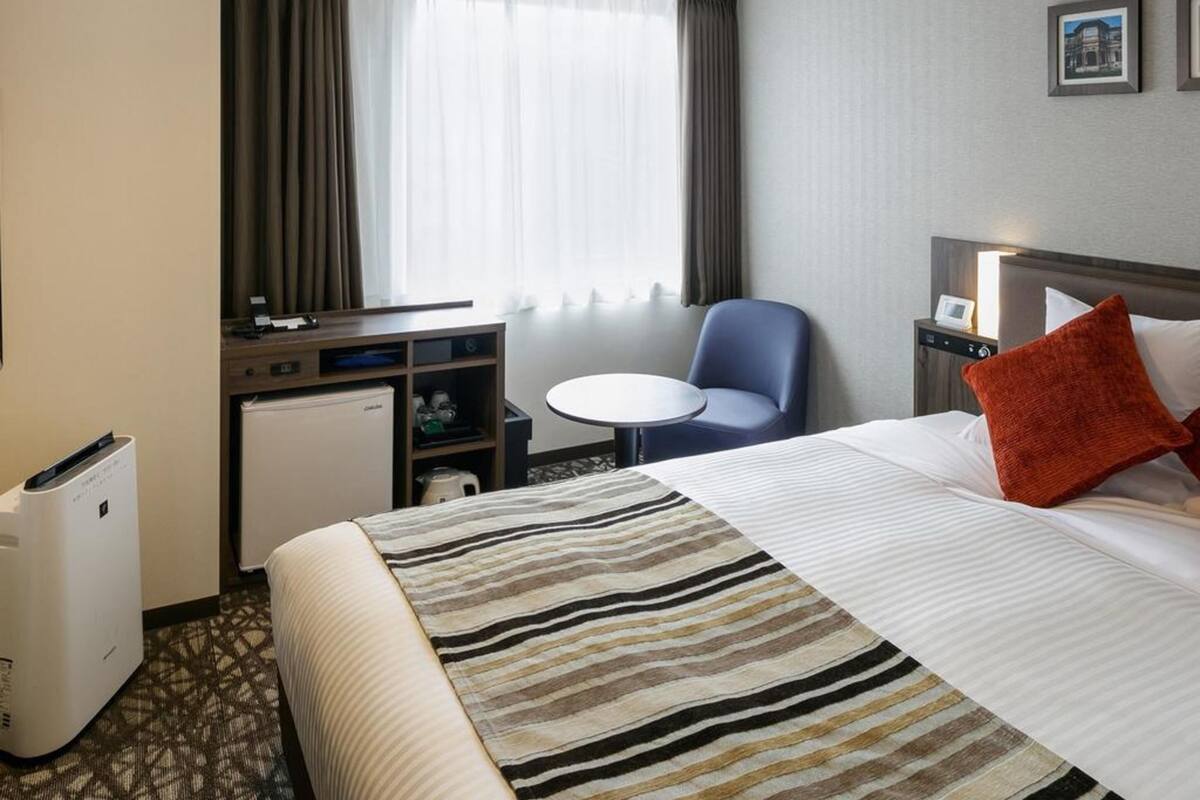 A hotel room with a bed, a desk, a chair, a small table, a refrigerator, and a window with curtains.