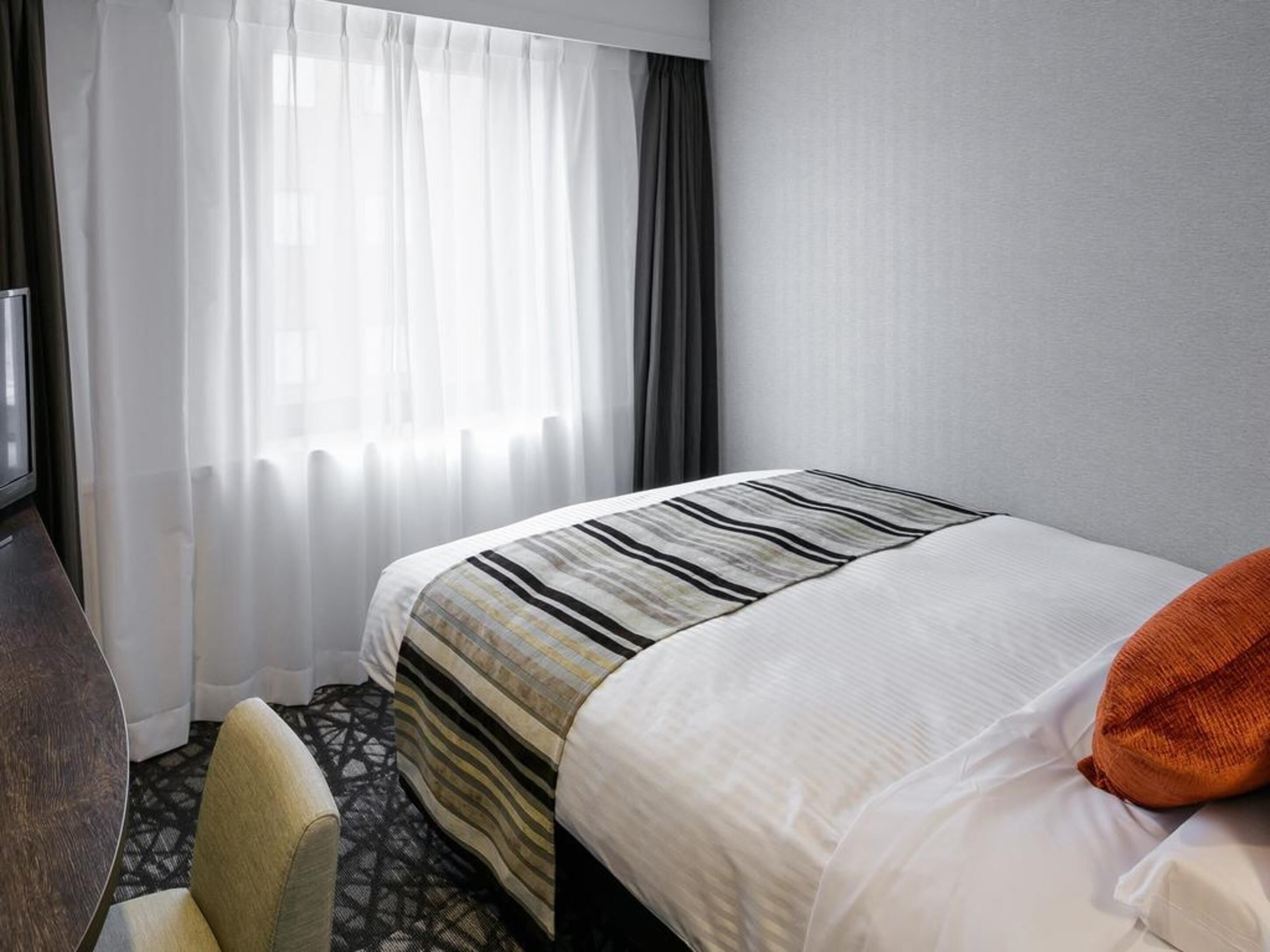 A hotel room with a bed, a chair, a TV, and a window with curtains.