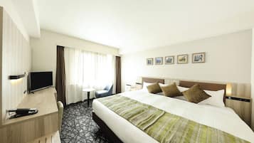 A hotel room with a bed, a desk with a TV, a chair, and a window with curtains.