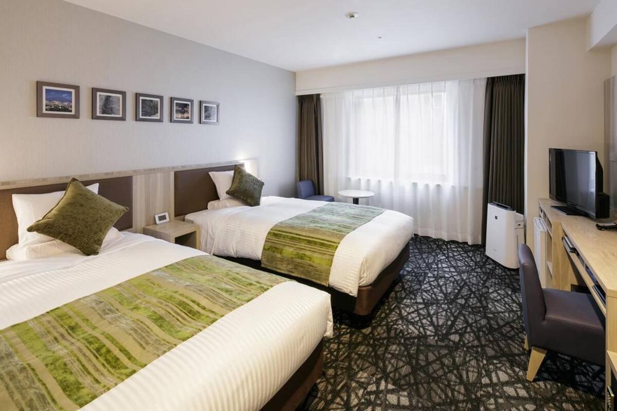 A hotel room with two beds, a desk, a TV, and a window with curtains.