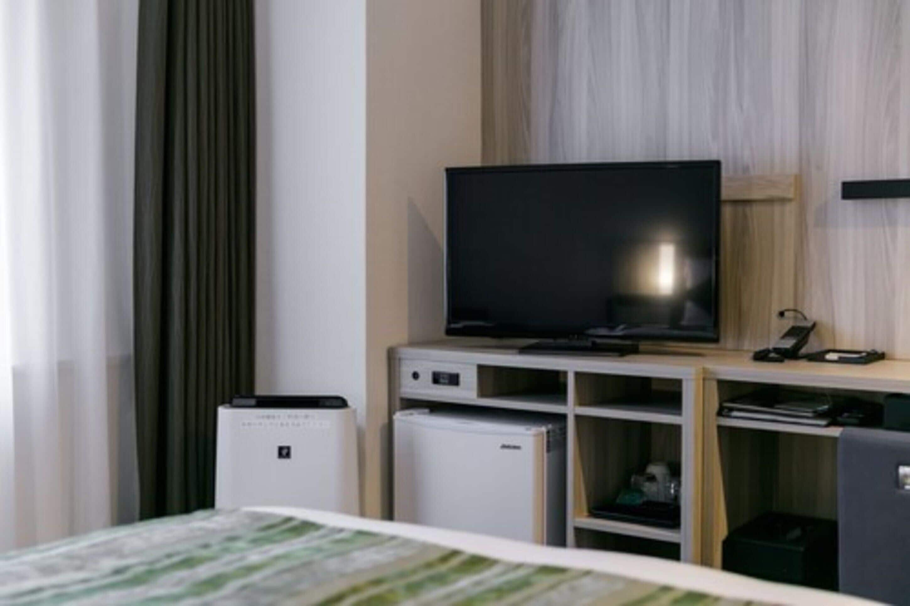 A modern hotel room with a flat-screen TV on a wooden stand, a small refrigerator, a bedside table, and a bed with a striped bedspread.