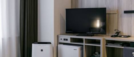 A modern hotel room with a flat-screen TV on a wooden stand, a small refrigerator, a bedside table, and a bed with a striped bedspread.