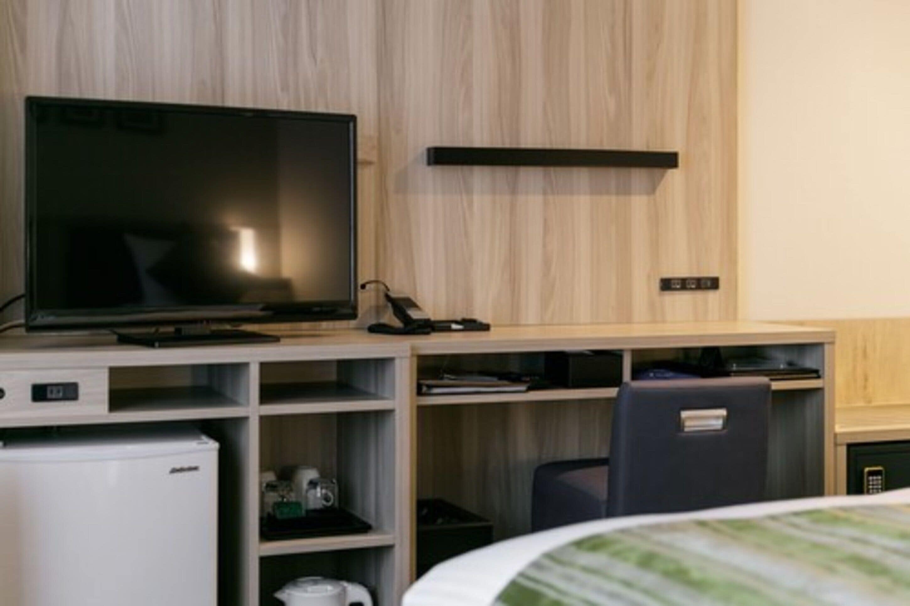 Une chambre d’hôtel équipée d’un téléviseur à écran plat, d’un bureau avec un ordinateur, d’un petit réfrigérateur et d’une chaise.