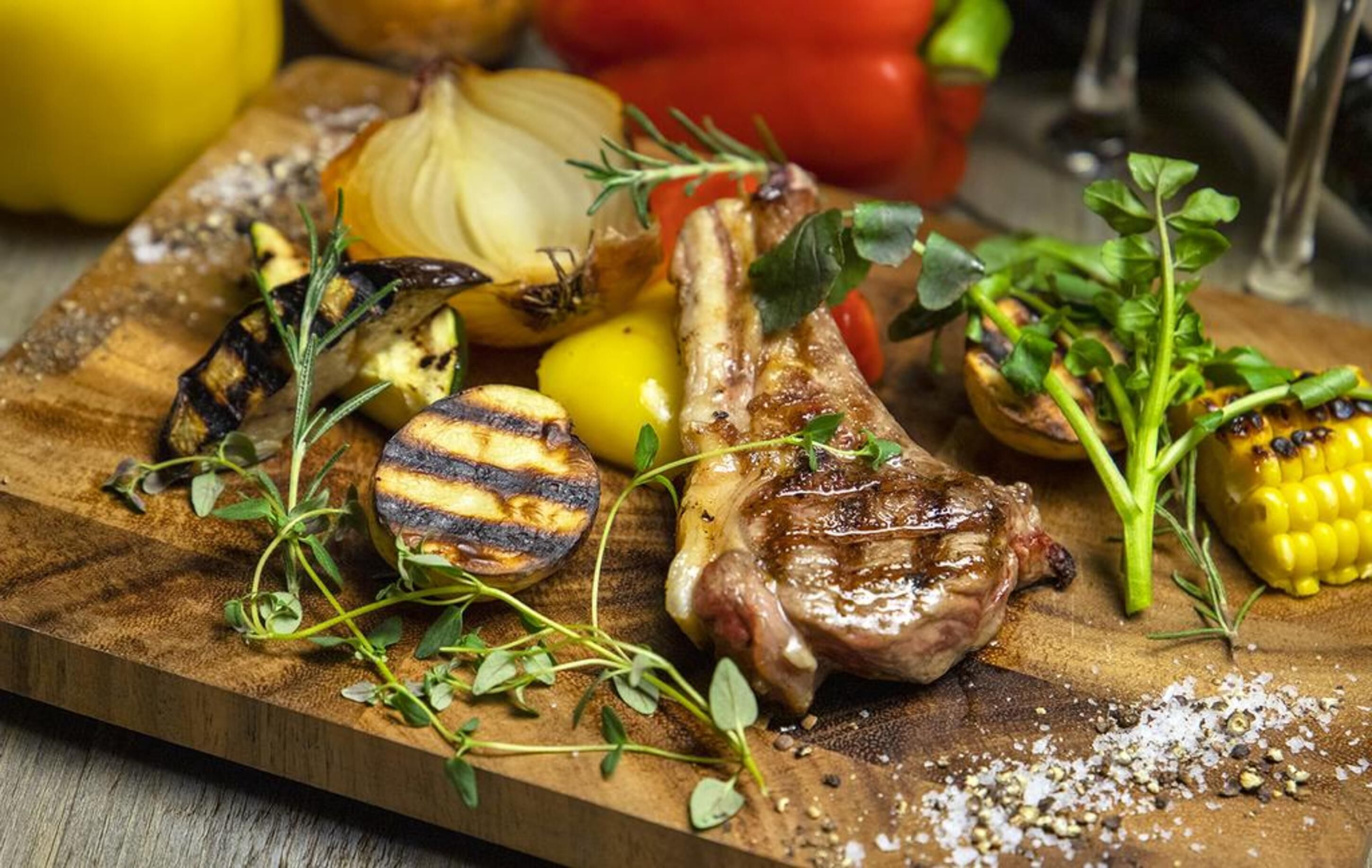 Ein Holzschnittbrett mit gegrilltem Gemüse, einem gegrillten Lammkotelett und Kräutern.