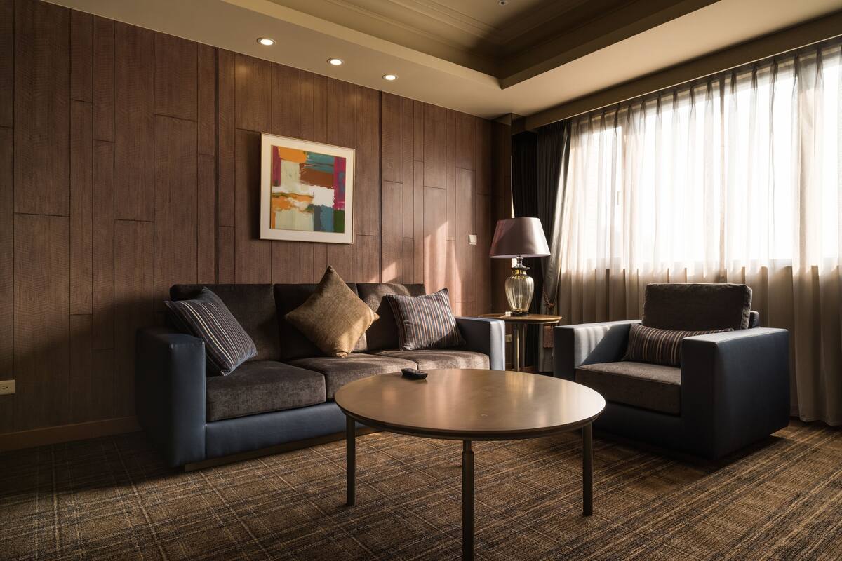 Una sala de estar moderna con un revestimiento de madera oscura, un sofá azul, un sillón más pequeño, una mesa de centro redonda y una lámpara sobre una mesita auxiliar.