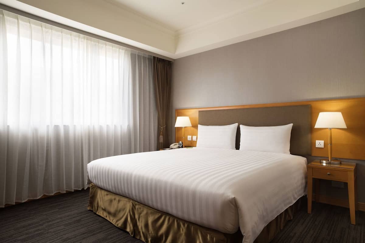 Habitación de hotel con una cama grande, dos lámparas de noche, una mesita de noche y una ventana con cortinas traslúcidas.