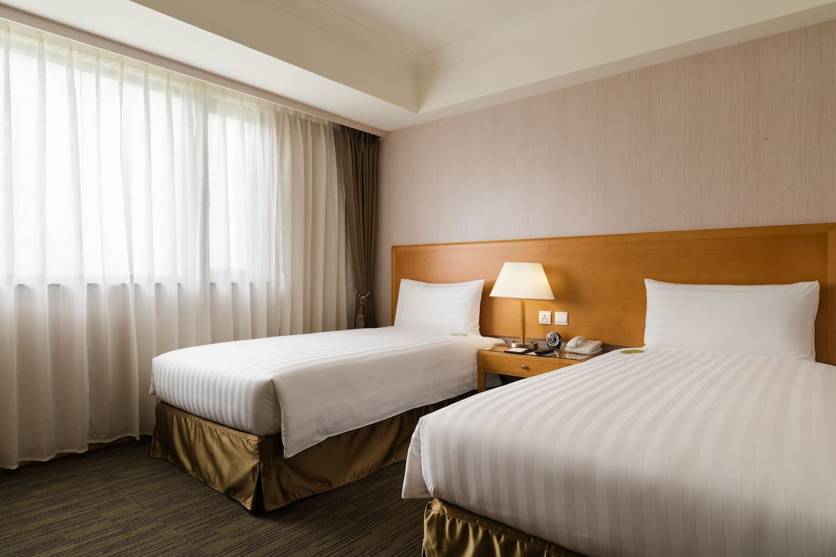 Una habitación de hotel con dos camas, una mesita de noche con lámpara y una ventana con cortinas.