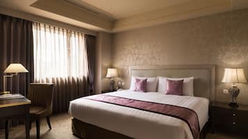 A hotel room with a large bed, two bedside tables with lamps, a desk with a chair, and a window with curtains.