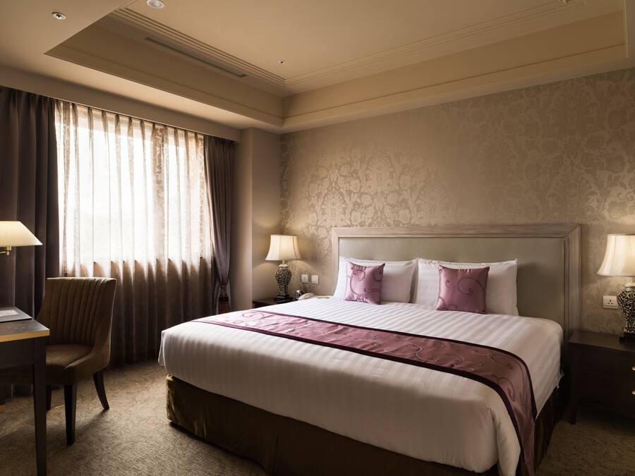 A hotel room with a large bed, two bedside tables with lamps, a desk with a chair, and a window with curtains.