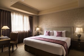 Habitación de hotel con una cama grande, dos mesitas de noche con lámparas, un escritorio con una silla y una ventana con cortinas.
