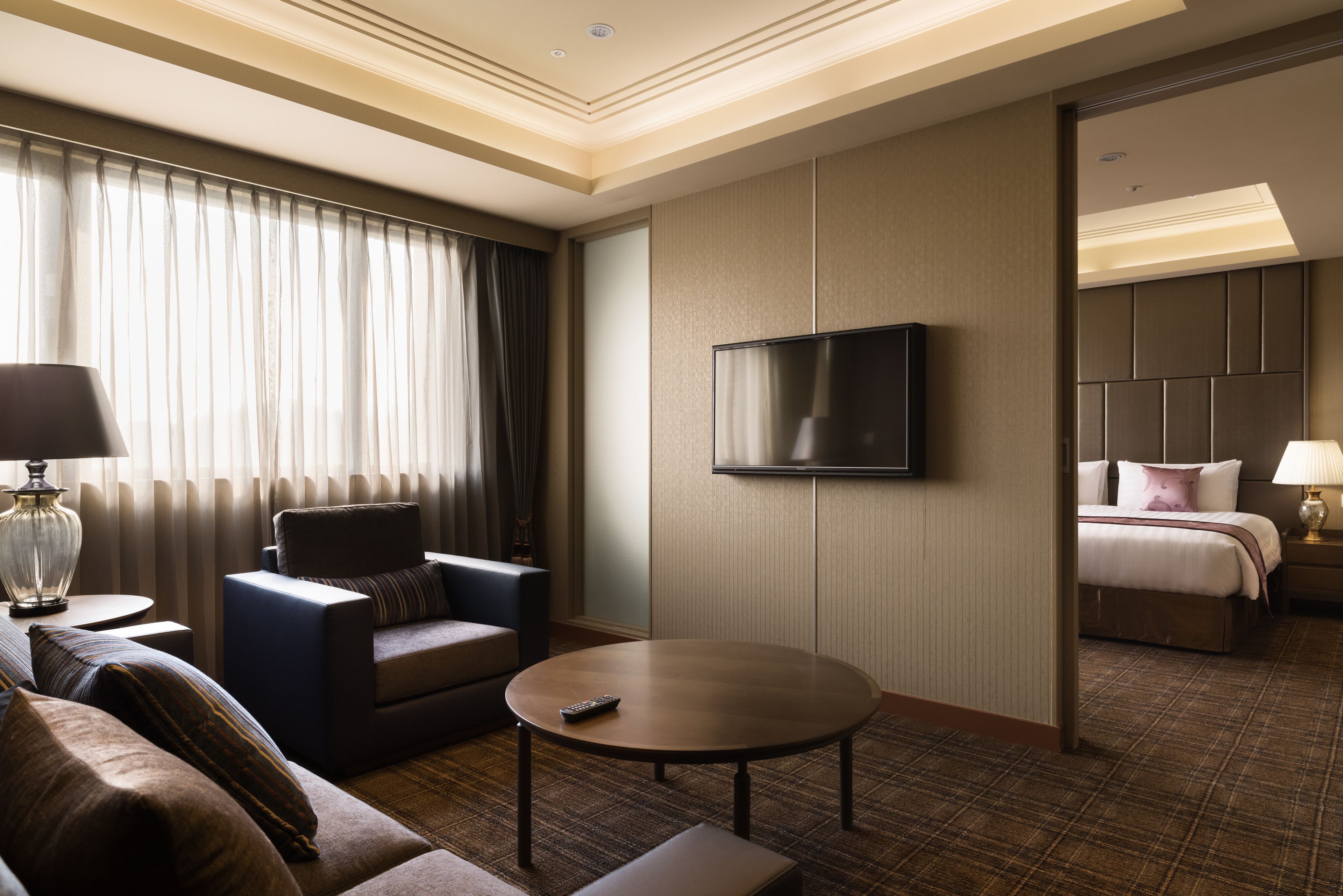Habitación de hotel con sofá, sillón, una mesa de centro redonda, televisor y una cama con mesita de noche.