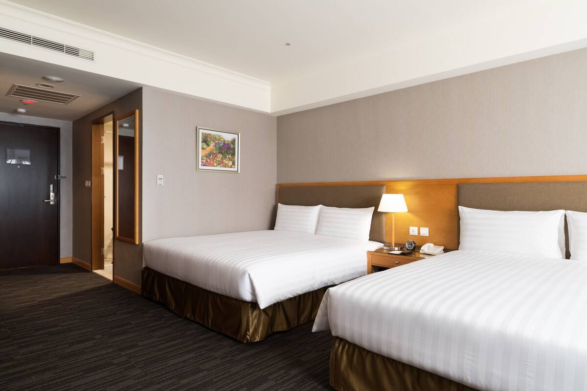 Habitación de hotel con dos camas, una mesita de noche, una lámpara y una puerta que da a otra habitación.