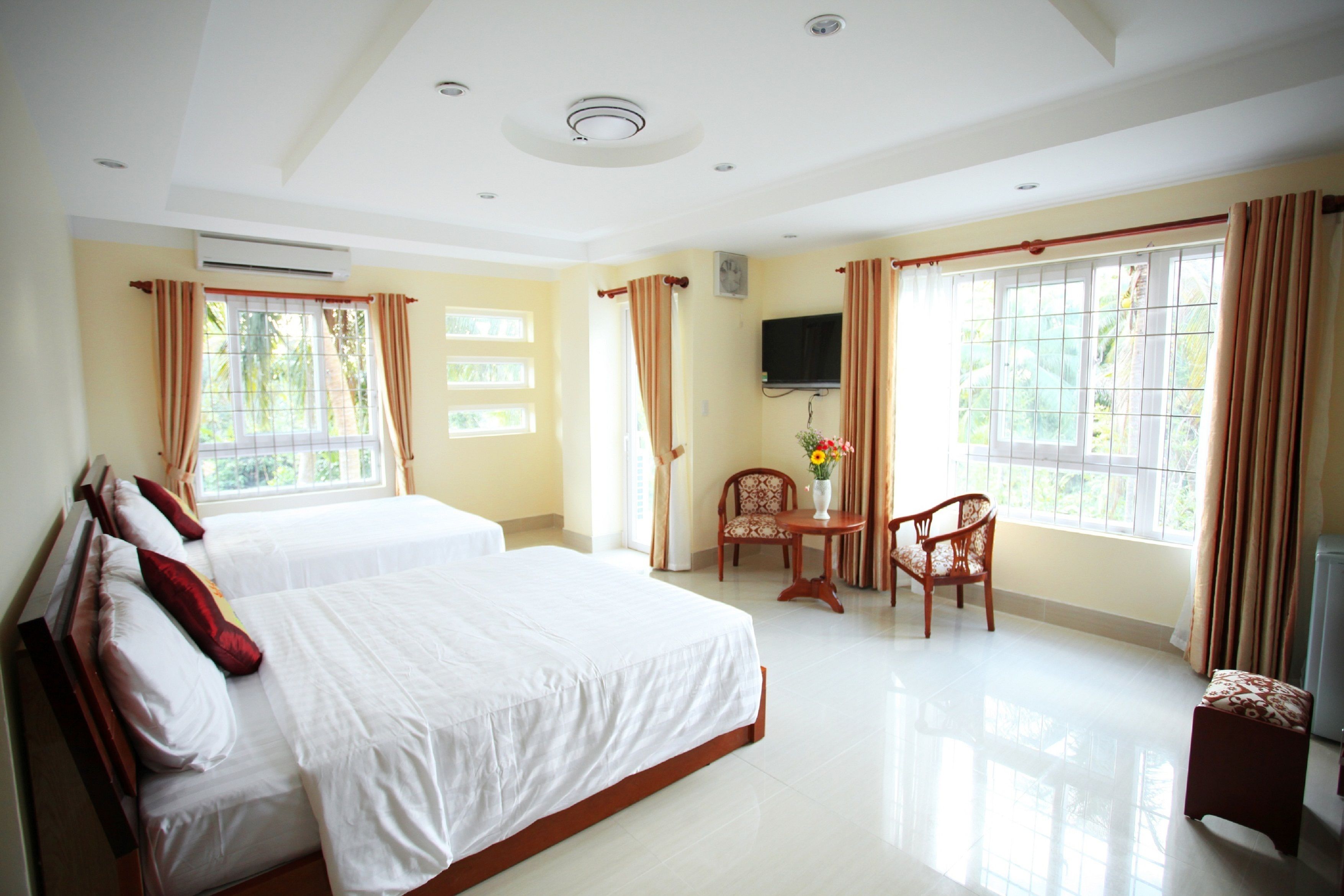 A hotel room with two beds, a TV, a small table with a vase, and large windows with curtains.