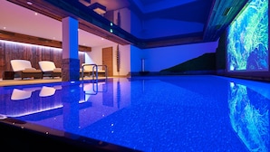 A modern indoor swimming pool with a blue-lit water feature, lounge chairs, and a large aquarium display. - Boutique Hotel Herzhof Garni - Apartments (Mittelberg)