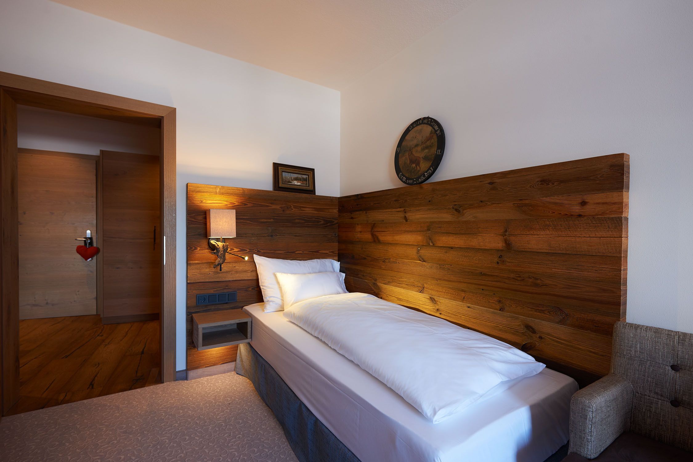 A bedroom with a wooden headboard, a bed with white linens, a bedside table with a lamp, and a small picture frame on the wall.