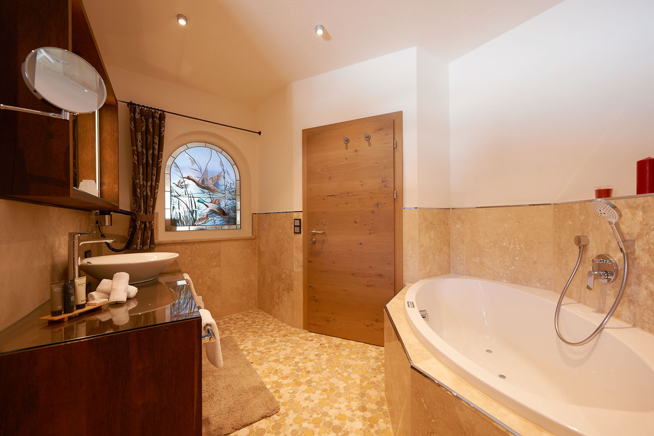 A modern bathroom with a wooden door, a large bathtub, a vanity with a mirror, and a window with a view of trees.