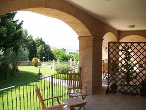 Un balcón con banca de madera y una celosía, con vista a un jardín cuidado y un edificio distante.