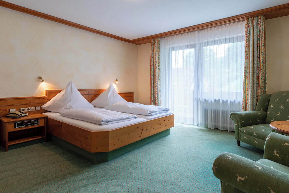 Quarto de hotel com cama de madeira, duas mesas de cabeceira, uma poltrona verde, uma mesa redonda pequena e uma janela grande com cortinas finas.