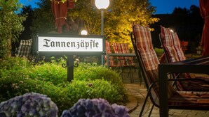 Un’area giardino con un cartello che recita “Tannenjapfle”, circondata da vegetazione lussureggiante e un’area salotto all’aperto.