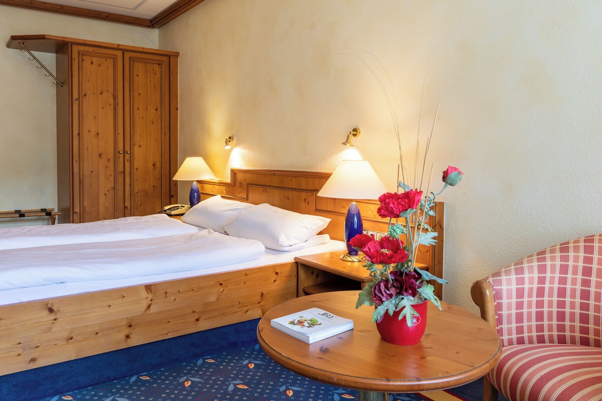 Quarto de hotel com uma cama de casal, duas luminárias de cabeceira, uma mesinha de centro com um livro e um vaso de flores, uma cadeira e armários de madeira.