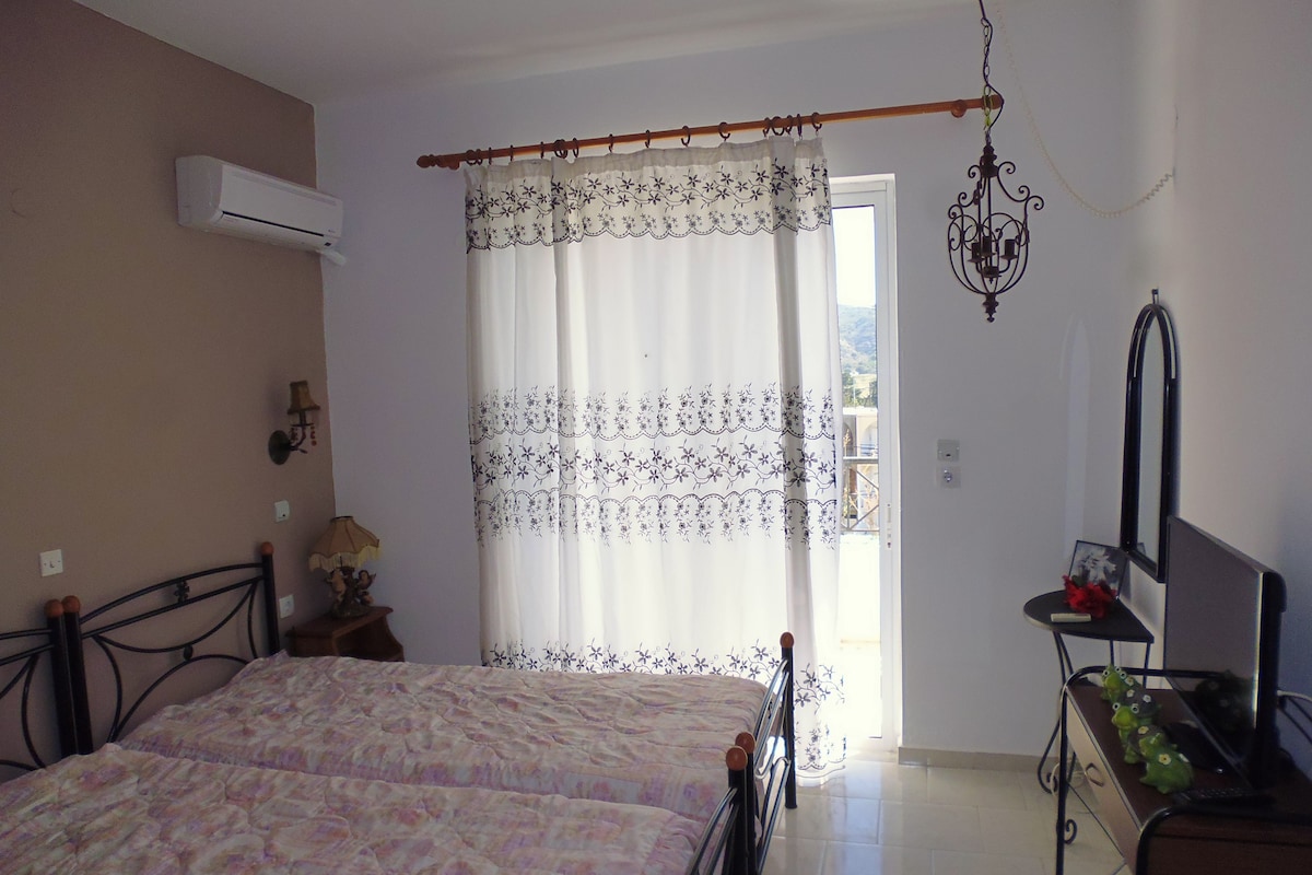 A bedroom with a bed, an air conditioner, a curtain, a bedside table, a lamp, a mirror, and a desk with a computer.