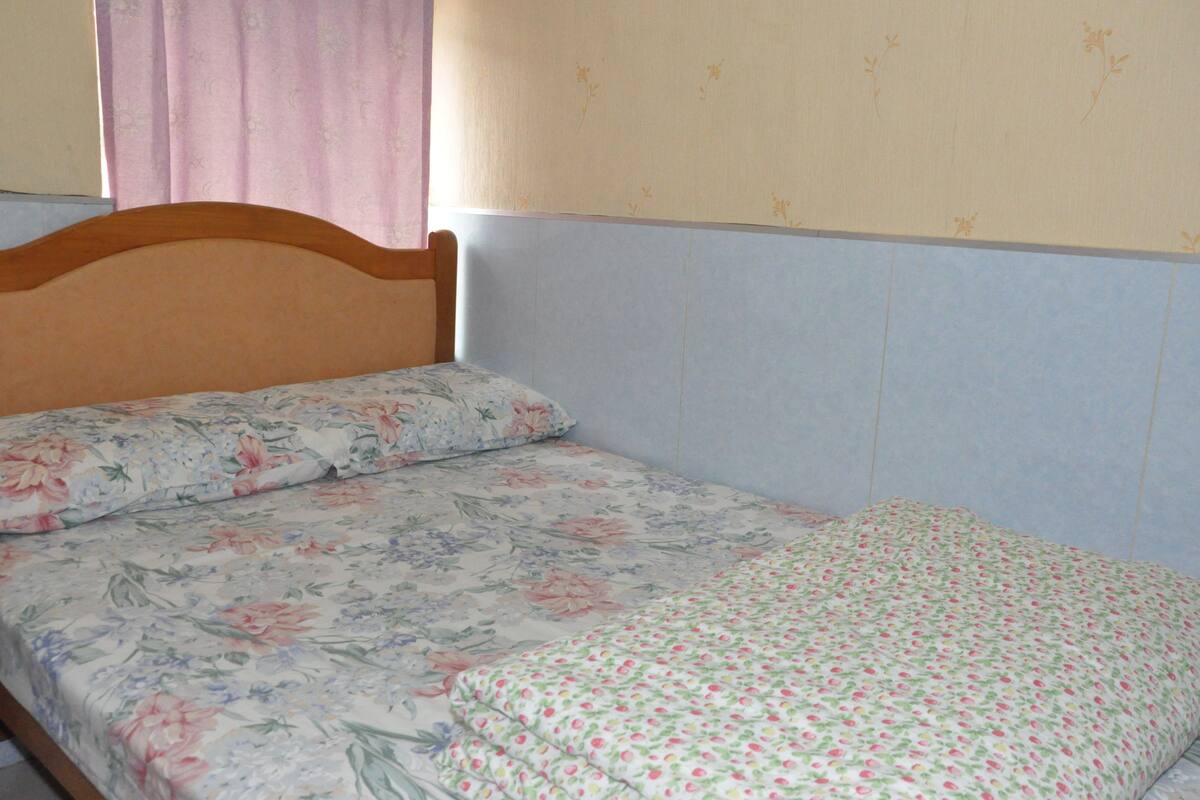 Una cama individual con ropa de cama floral y cabecera de madera en una habitación con paredes azul claro y una cortina rosa.