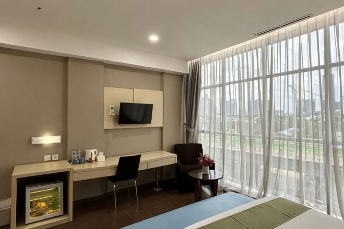 Une chambre d’hôtel comprenant un lit, un bureau avec une chaise, un téléviseur fixé au mur et une grande fenêtre avec des rideaux légers.
