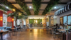 Een ruim café met houten vloer, zwarte stoelen en tafels. Er hangen planten en aan één kant is een bakstenen muur.