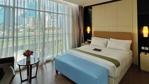Hotelkamer met een groot bed, een klein tafeltje met een bloevas en uitzicht op de stad door grote ramen.