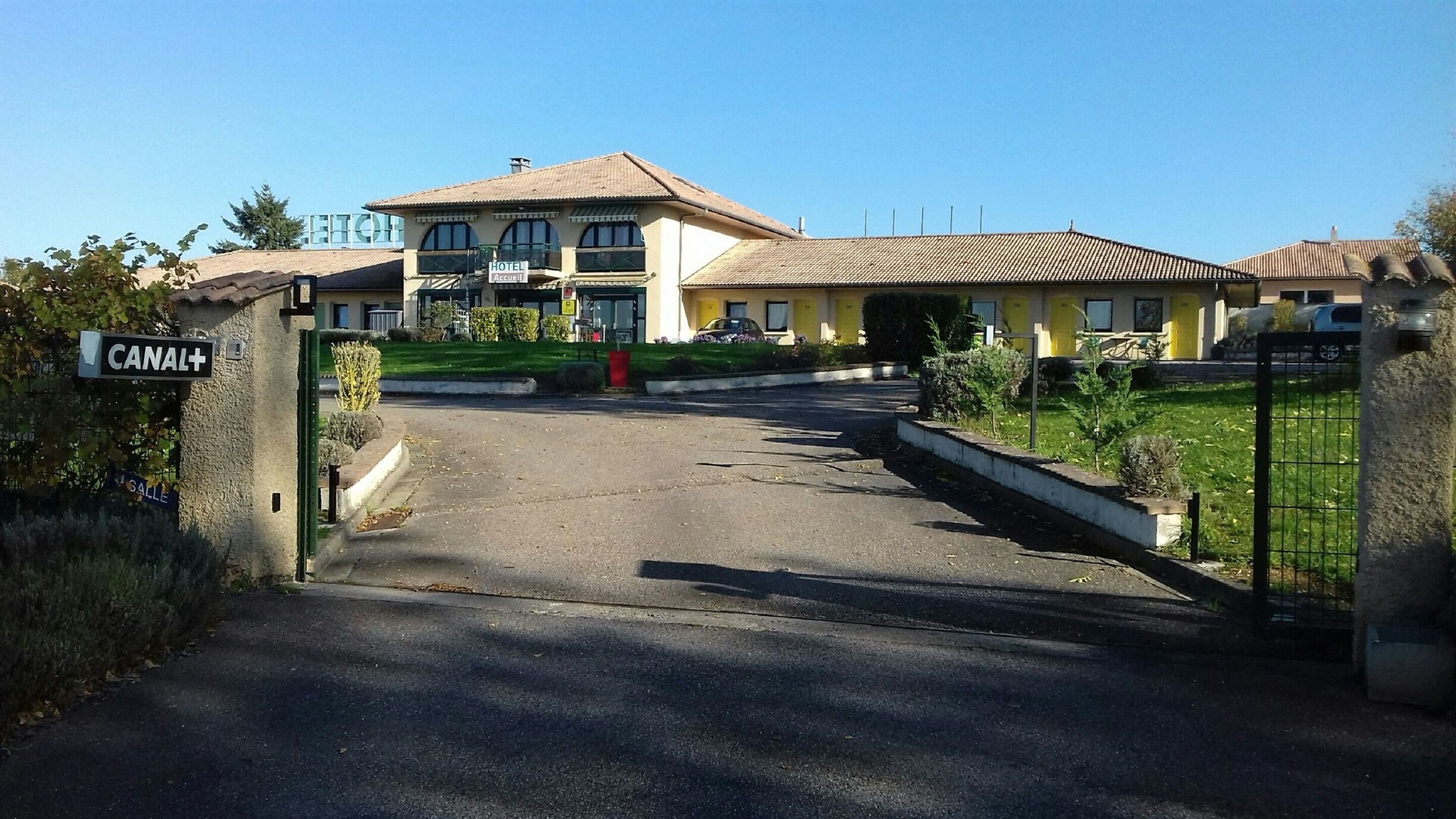 En hotellentrén med en asfalterad uppfart som leder fram till en byggnad med en gul fasad och en skylt med texten ”HOTEL Accueil”.