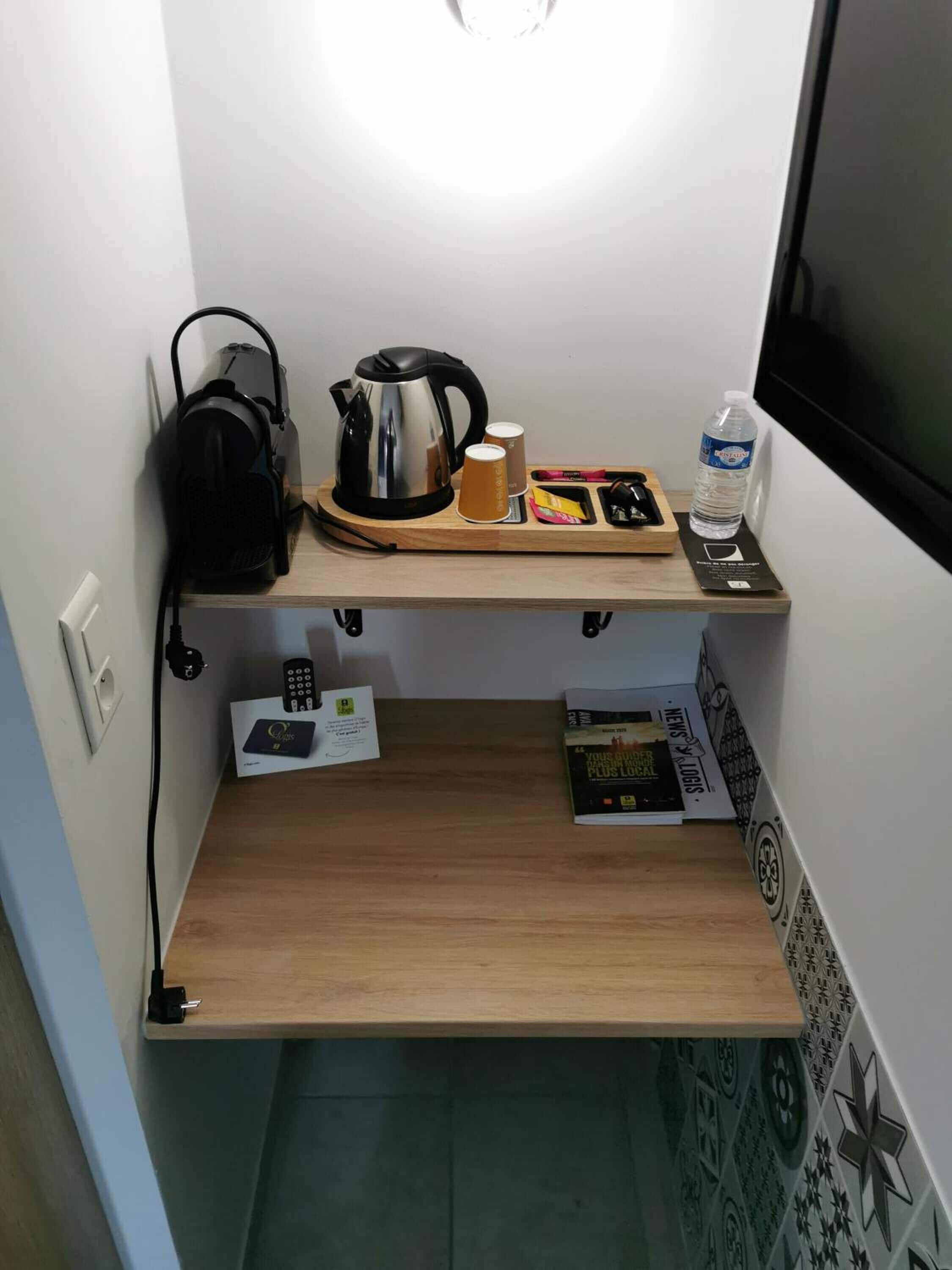 Een kleine kitchenette met een koffiezetapparaat, waterkoker, kopjes, een waterfles en een afstandsbediening op een houten plank.