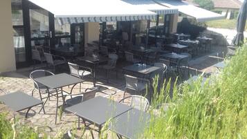 Eine Terrasse im Freien mit mehreren Tischen und Stühlen, von denen einige mit Sonnenschirmen beschattet werden, und einem gepflasterten Boden.