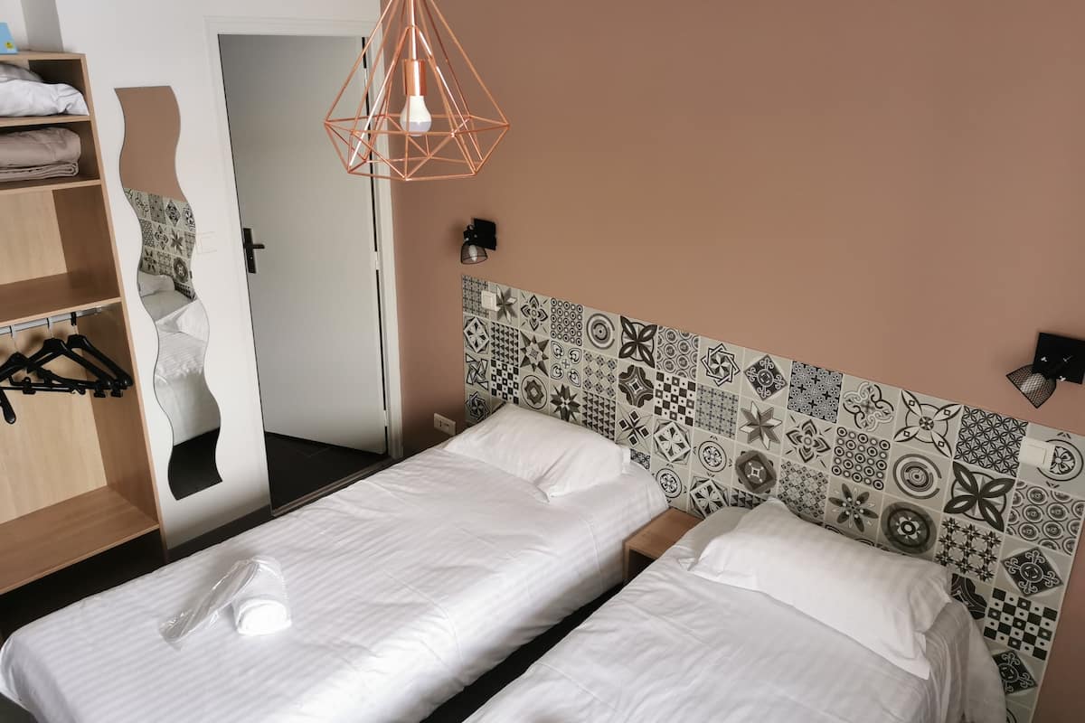 Quarto de hotel com duas camas, um guarda-roupa de madeira, um candeeiro pendente e uma cabeceira estampada.