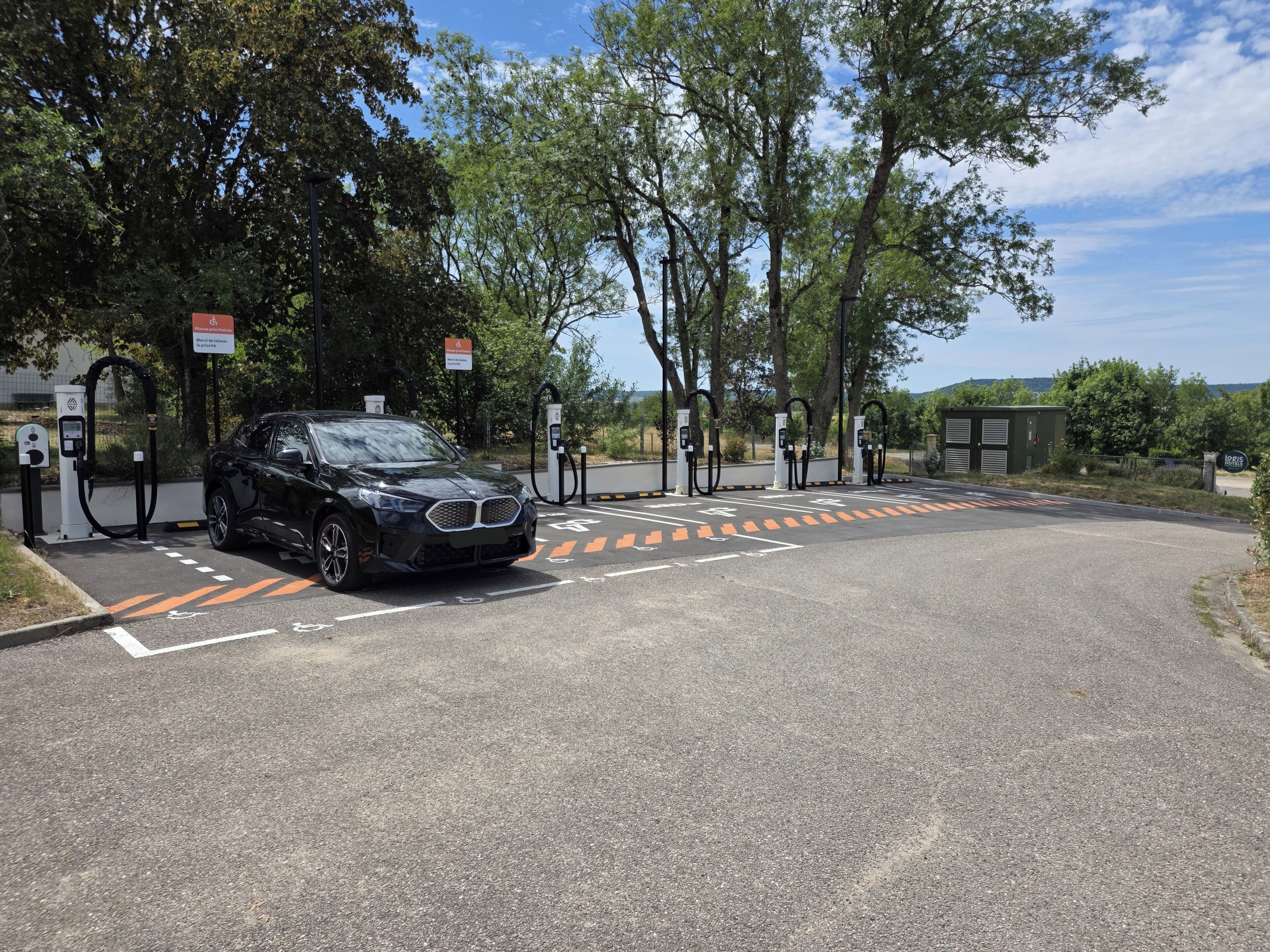 A black car parked at a charging station with trees and a clear sky in the background.
