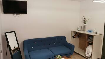 Een moderne woonkamer met een blauwe bank, een klein houten bijzettafeltje, een flatscreen televisie aan de muur en een inbouwkast met een kapstok.