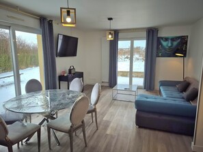 A modern living room with a glass dining table, a blue sofa, and a flat-screen TV mounted on the wall. - Hotel La Haie des Vignes (Allain)