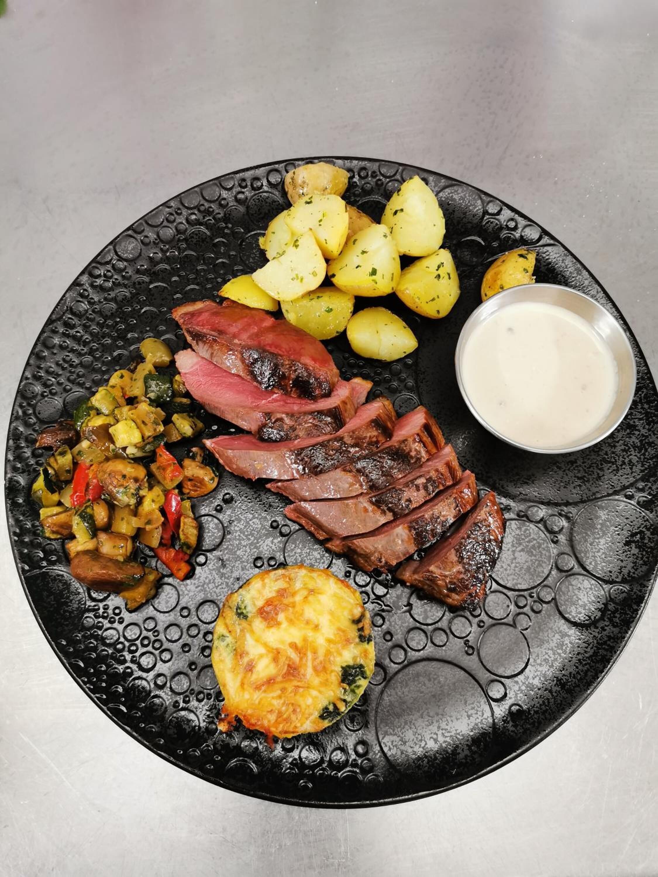 A plate with sliced meat, roasted potatoes, a vegetable medley, and a small bowl of sauce.