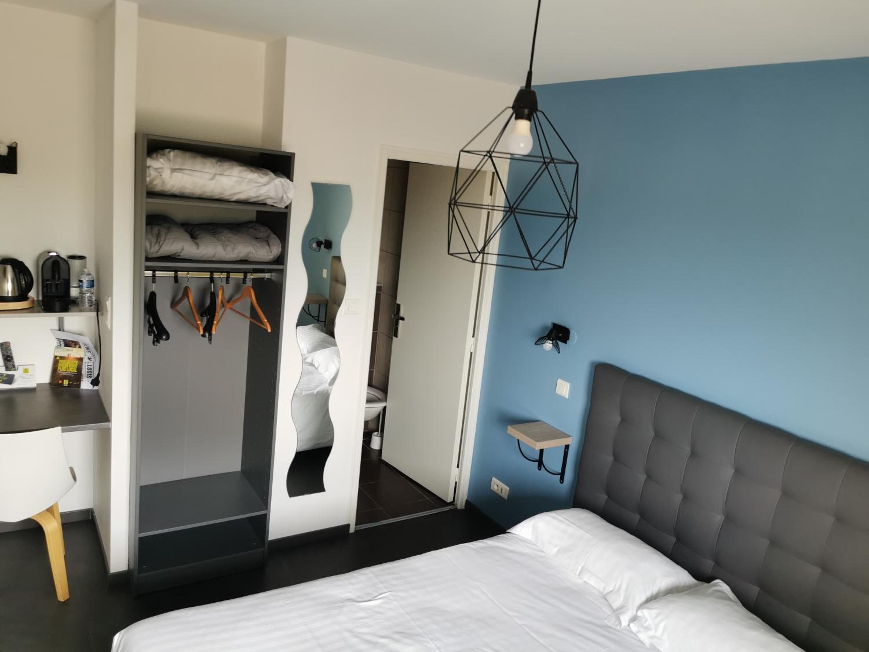 A modern hotel room with a bed, a desk, a wardrobe, and a bathroom visible through an open door.