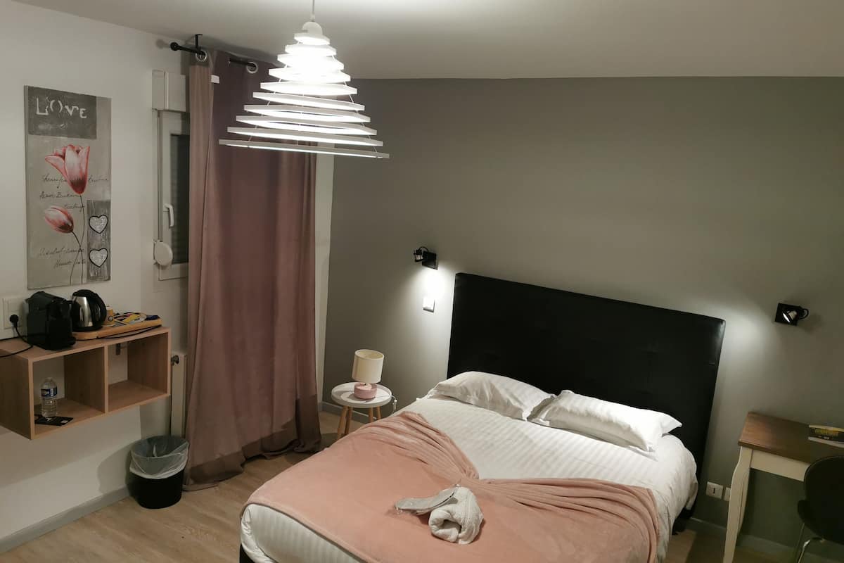 Quarto de hotel com cama, mesa de cabeceira, luminária, aquecedor fixado na parede, prateleira com artigos de higiene pessoal e um objeto decorativo na parede.