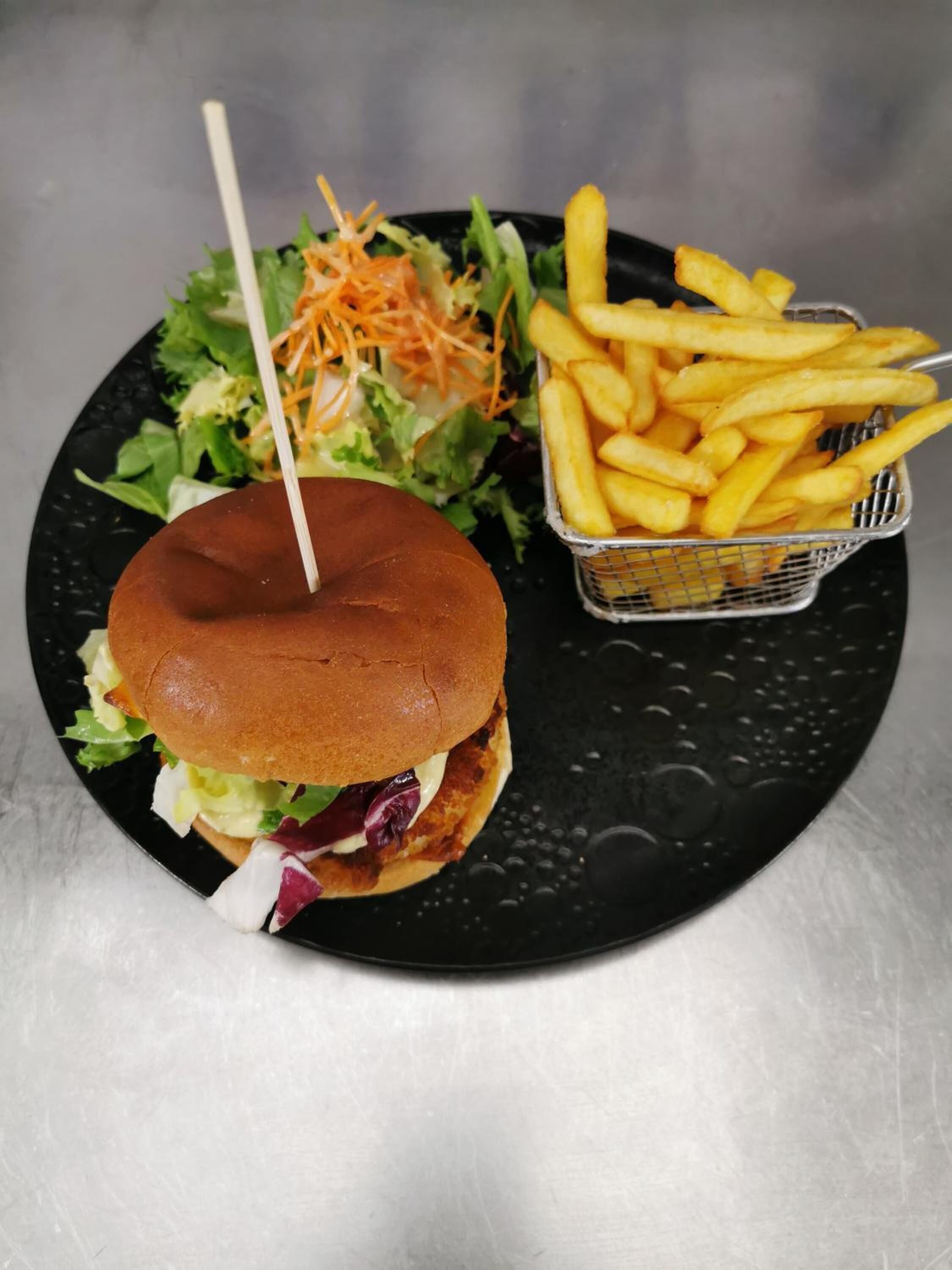 Una hamburguesa con un palillo, una ensalada pequeña y una cesta de papas fritas sobre un plato negro.